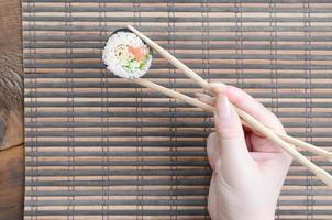 Eine Hand mit Essstäbchen hält eine Sushi-Rolle auf einem Hintergrund einer Bambusstroh-Sewing-Matte. traditionelles asiatisches essen foto