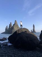 Mann steht auf einem Felsen am schwarzen Strand von Reynisfjara, Island, mit Wind im Haar foto