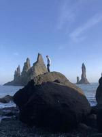 Mann steht auf einem Felsen am schwarzen Strand von Reynisfjara, Island, mit Wind im Haar foto