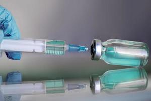 Fläschchen gefüllt mit flüssigem Impfstoff im medizinischen Labor mit Spritze. Die Hand des Arztes hält Ampulle und Spritze auf der Glasoberfläche