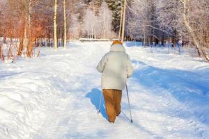 Erwachsene Frau in Wintersportbekleidung mit Stöcken für Nordic Walking auf Winterlandschaft Stadtpark Hintergrund, Rückansicht. reife frau, die draußen übung macht. winter soprt gesundes lebensstilkonzept foto