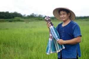 asiatischer Bauer ist auf Reisfeld, trägt Hut, blaues Hemd, hält thailändischen Lendenschurzschal, um Schweiß während der Arbeit abzuwischen. konzept, landwirtschaftsberuf. ländlicher Lebensstil. hart arbeitend. foto
