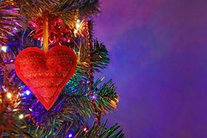 Rotes Herz, handgefertigte Weihnachtsdekoration mit Ornament auf Tanne in der Feiertagsnacht, bunte Bokeh-Girlande, rote Perlen, unscharfer dunkelvioletter Hintergrund. weihnachtsgrußkarte mit kopienraum. foto