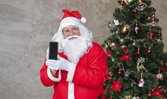 der weihnachtsmann feiert weihnachten in freude und aufregung, während er sein handy neben dem voll geschmückten weihnachtsbaum für verkaufsförderung und online-shopping-konzept hält foto