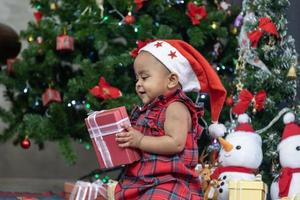 afroamerikanisches baby lächelt glücklich, während es kleine geschenkboxen von den eltern erhält, während es sich in weihnachtskleid und weihnachtsmütze mit weihnachtsbaum auf dem rücken für die saisonfeier kleidet foto