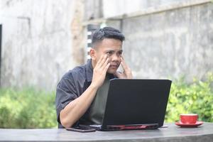 attraktiver junger asiatischer mann, der einen laptop verwendet, der im co-working space mit unglücklichem gesicht verwirrt ist foto