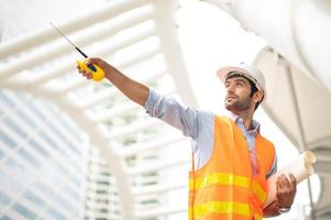 Kaukasischer Ingenieur benutzt ein Walkie-Talkie zum Reden, trägt eine orangefarbene Weste und einen großen Schutzhelm und hält mit der anderen Hand den weißen Grundriss in der Baustelle der Innenstadt. foto