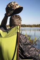 Der junge, nicht erkennbare Fischer fängt Fische, die auf einem Stuhl am Ufer eines Flusses oder Sees sitzen. Der Angler sitzt mit Angelausrüstung auf einem Stuhl. ein perfekter Urlaub am Fluss mit Angelrute. foto