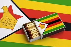 Simbabwes Flagge wird in einer offenen Streichholzschachtel gezeigt, die mit Streichhölzern gefüllt ist und auf einer großen Flagge liegt foto
