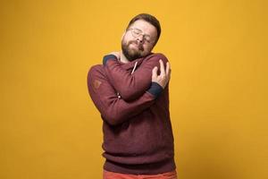 charmanter kaukasischer bärtiger mann mit brille umarmt sich zärtlich und schließt verträumt die augen. gelber Hintergrund. foto