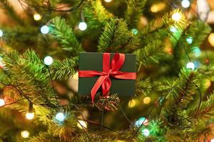 weihnachtsgeschenk am weihnachtsbaum mit girlandenlichtern, geschenk in grüner schachtel mit roter schleife, neujahrsferien vorhanden foto