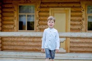 Junge geht in der Nähe des Hauses. Kind auf der Bühne. Junge auf dem Hintergrund eines Holzhauses. Junge im weißen Hemd foto