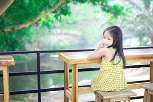 kleines asiatisches Mädchen, das auf dem Stuhl an einem Holztisch sitzt foto