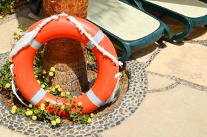 der orangefarbene rettungsring für die sicherheit beim schwimmen ruht auf einem steinboden vor einem hintergrund aus gelben blumen und sonnenliegen am tropischen marinen exotischen südlichen wärmeresort foto