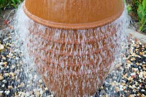 Ein schöner kleiner Brunnen in Form einer braunen Vase, ein Krug mit fallenden Wassertropfen auf farbigen Steinen, die in einem Blumenbeet mit grünen Pflanzen stehen foto