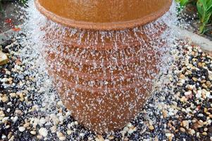 Ein schöner kleiner Brunnen in Form einer braunen Vase, ein Krug mit fallenden Wassertropfen auf farbigen Steinen, die in einem Blumenbeet mit grünen Pflanzen stehen foto