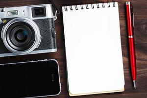 Klassische Kamera mit leerer Notizblockseite und rotem Stift auf dunkelbraunem Holztisch, Vintage-Tisch mit Telefon und grüner Blume foto