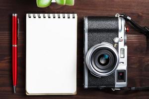 Klassische Kamera mit leerer Notizblockseite und rotem Stift auf dunkelbraunem Holztisch, Vintage-Tisch mit Telefon und grüner Blume foto