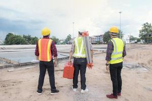 Rückansicht Gruppe von Ingenieuren, Technikern und Architekten mit Schutzhelmplanung über Bauplan mit Blaupause im Hintergrund des modernen Stadtbaus, Baustelle, Industrie und Arbeiterkonzept foto