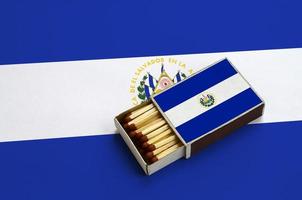 Die el-Salvador-Flagge wird in einer offenen Streichholzschachtel gezeigt, die mit Streichhölzern gefüllt ist und auf einer großen Fahne liegt foto