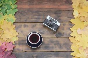 Eine Tasse Tee und eine alte Kamera inmitten einer Reihe von vergilbten gefallenen Herbstblättern auf einer Hintergrundfläche aus natürlichen Holzbrettern von dunkelbrauner Farbe foto