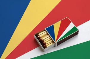 Die Flagge der Seychellen wird in einer offenen Streichholzschachtel gezeigt, die mit Streichhölzern gefüllt ist und auf einer großen Flagge liegt foto
