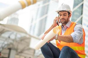 Kaukasischer Ingenieur benutzt ein Smartphone zum Reden, trägt eine orangefarbene Weste und einen großen Schutzhelm und hält in der anderen Hand den weißen Grundriss in der Baustelle der Innenstadt. foto