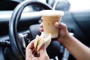 asiatische fahrerin hält eiskaffeetasse und sandwichbrot zum essen und trinken im auto, gefährlich und riskiert einen unfall. foto