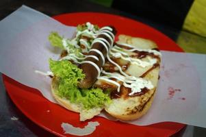 indonesischer lokaler hotdog. Roastbeef, Wurst, Sesambrot, Salat, Mayonaise und Sauce. Hotdog-Herstellungsprozess. Straßenessen. foto