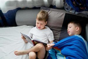 Der kleine Bruder liest ein Buch auf dem Bett für einen Ältesten. Freundschaft zwischen Brüdern. Liebe zum Lesen. Lerche und Eule unter den Menschen. 2 Jungen auf dem Bett lesen ein Buch. Gute-Nacht-Geschichten foto