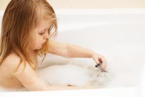 Kleines Mädchen mit blonden Haaren, das mit Schaum in einer Badewanne spielt. Mädchen nimmt ein Bad. Mädchen spielt mit Spielzeug im Bad foto