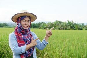 asiatischer frauenbauer ist auf landwirtschaftsland, trägt hut, thailändischen lendenschurz bedeckten kopf, blaues hemd und macht handgeste, um etwas zu präsentieren. konzept, landwirtschaftsberuf. Bauer mit Bio-Reis. foto