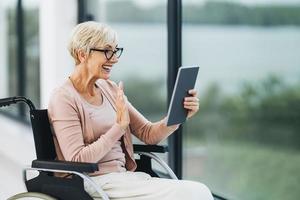 Behinderte Seniorin, die am Fenster sitzt und ein digitales Tablet verwendet foto