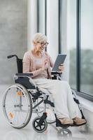Behinderte Seniorin, die am Fenster sitzt und ein digitales Tablet verwendet foto