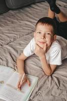 Porträt eines Jungen, der Hausaufgaben macht und auf einem grauen Sofa liegt. Ein Teenager schreibt in ein Arbeitsbuch auf der Couch. Homeschooling eines 9-jährigen Jungen. freies Lernen foto