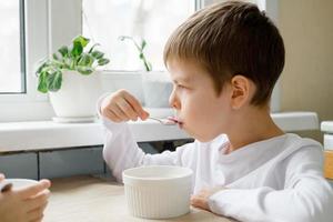 Kind isst farbiges Müsli am Tisch in einer weißen Küche. Jungenfrühstück. Frühstücksflocken vor der Schule. ein schneller Imbiss eines Schuljungen am Morgen. foto