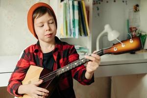 Junge mit rotem Hut und kariertem Hemd spielt Balalaika. hübscher junge, der seine gitarre hält. Musikunterricht zu Hause. Hobby für die Seele. Musikunterricht zu Hause foto