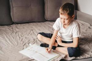 Junge macht Hausaufgaben, während er auf dem grauen Sofa sitzt. ein kaukasischer teenager schreibt in ein arbeitsbuch auf der couch. Homeschooling eines 9-jährigen Jungen. freies Lernen foto