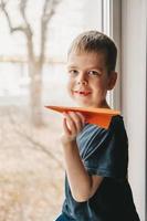 ein Junge mit einem orangefarbenen Papierflieger sitzt am Fenster und schaut auf die Straße hinaus. Der Junge träumt vom Reisen. geschlossene Grenzen foto