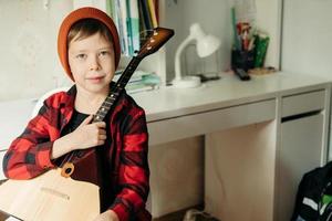 Junge mit rotem Hut und kariertem Hemd spielt Balalaika. hübscher junge, der seine gitarre hält. Musikunterricht zu Hause. Hobby für die Seele. Musikunterricht zu Hause foto