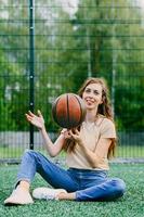 lächelndes mädchen mit braunen langen haaren sitzt mit einem basketball in den händen auf dem rasen. Mädchen schaut in den Rahmen. schöner Basketballspieler sitzt mit dem Ball auf dem Rasen. Mädchen treibt Sport. foto