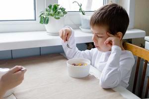 Kind isst farbiges Müsli am Tisch in einer weißen Küche. Jungenfrühstück. Frühstücksflocken vor der Schule. ein schneller Imbiss eines Schuljungen am Morgen. foto