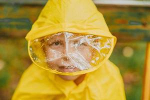 Porträt eines Jungen in einem gelben Regenmantel auf der Straße während des Regens. Regenhaubenschutz. Regentropfen auf einer transparenten Kapuze, die das Gesicht bedeckt. helle Kleidung für Kinder für einen Spaziergang. foto