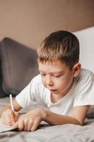 Porträt eines Jungen, der Hausaufgaben macht und auf einem grauen Sofa liegt. Ein Teenager schreibt in ein Arbeitsbuch auf der Couch. Homeschooling eines 9-jährigen Jungen. freies Lernen foto