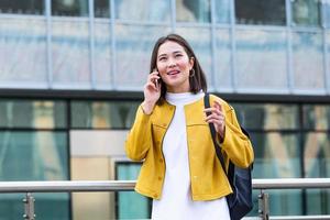 asiatische fröhliche junge frau, die die straße entlang geht und telefoniert. asiatische frau, die auf der straße geht, süßes trendiges outfit trägt und mit ihrem smartphone spricht. foto