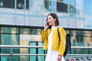 asiatische fröhliche junge frau, die die straße entlang geht und telefoniert. asiatische frau, die auf der straße geht, süßes trendiges outfit trägt und mit ihrem smartphone spricht. foto