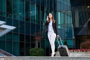 Geschäftsfrau, die mit Koffern auf Geschäftsreise geht, während sie mit ihrem Handy spricht. Geschäftsfrau am internationalen Flughafen, die für eine Flugreise zum Terminaltor umzieht foto