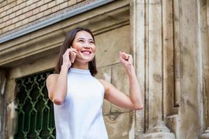 asiatische fröhliche junge frau, die die straße entlang geht und telefoniert. asiatische glückliche frau, die auf der straße geht, süßes trendiges outfit trägt und mit ihrem smartphone spricht. foto