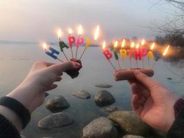 Brennende Inschrift zum Geburtstag aus Feiertagskerzen in den Händen eines Mannes und einer Frau gegenüber dem Wasser des Ozeanseeflusses. konzept geburtstagsfeier in der natur, im freien foto