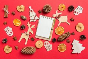 Draufsicht auf Notizbuch, roter Hintergrund mit festlichen Spielsachen und Weihnachtssymbolen, Rentieren und Neujahrsbäumen. Urlaubskonzept foto